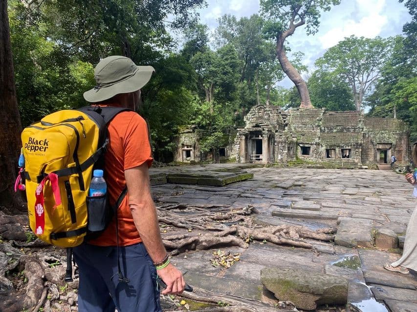Un viaggiatore con un cappello e uno zaino giallo osserva antiche rovine di templi in pietra invase da alberi e radici della giungla.