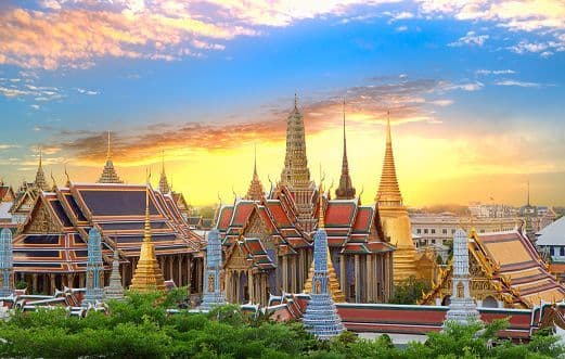 An ornate temple complex with golden spires and tiered roofs rises above lush green trees during a vibrant sunset.