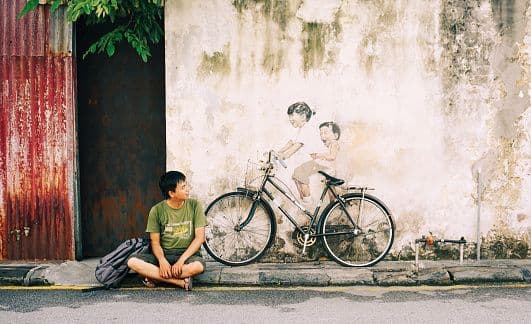 A man sits cross-legged on a sidewalk next to a real bicycle incorporated into a wall mural of two children riding it.