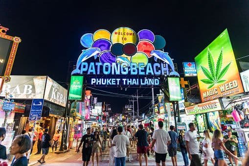 A crowd of people walking down a busy street at night, illuminated by a large colorful neon archway and various shop signs.