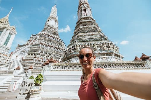 A smiling woman wearing sunglasses takes a selfie in front of two ornate white temple towers under a blue sky.