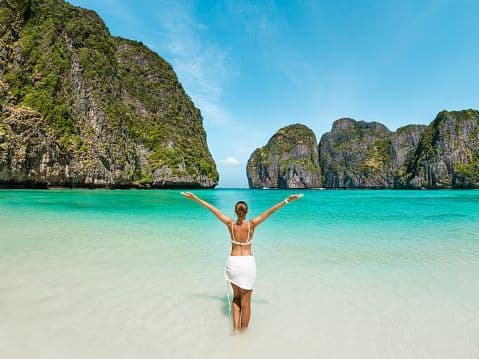 A woman in a white bikini stands with her arms outstretched in shallow turquoise water, facing large green rock formations in a bay.