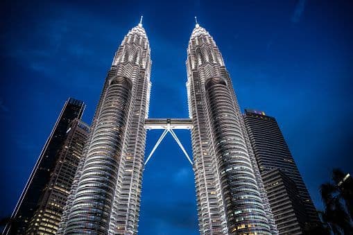 Two illuminated twin skyscrapers with a connecting skybridge, viewed from a low angle against a dark blue night sky.