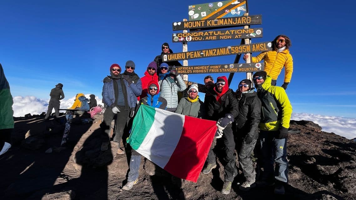 Un gruppo WeRoad in posa con una bandiera italiana davanti a un cartello di legno sulla cima di una montagna, molto al di sopra delle nuvole.