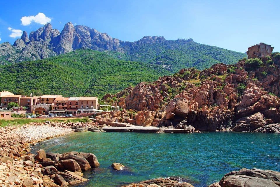 Ein Küstendorf, eingebettet in eine felsige Bucht mit türkisfarbenem Wasser, vor einer Kulisse steiler, grüner Berge unter blauem Himmel.