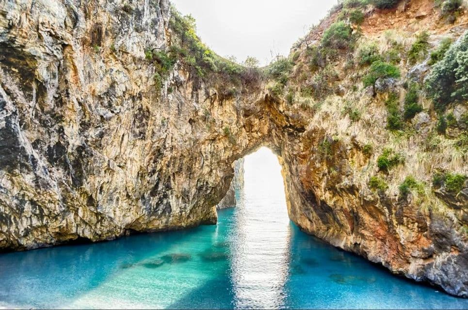 Un grande arco roccioso naturale si erge su acqua calma, limpida e turchese, con la luce del sole che filtra attraverso l'apertura.