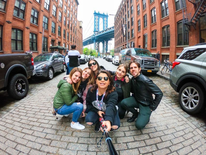 A WeRoad group trip takes a selfie on a cobblestone street with red brick buildings and a blue bridge in the background.