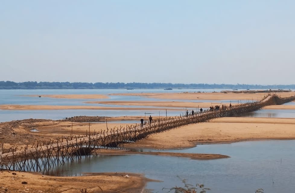 Un gruppo WeRoad attraversa un lungo ponte di bambù che si estende su un ampio fiume con grandi banchi di sabbia, sotto un cielo sereno.