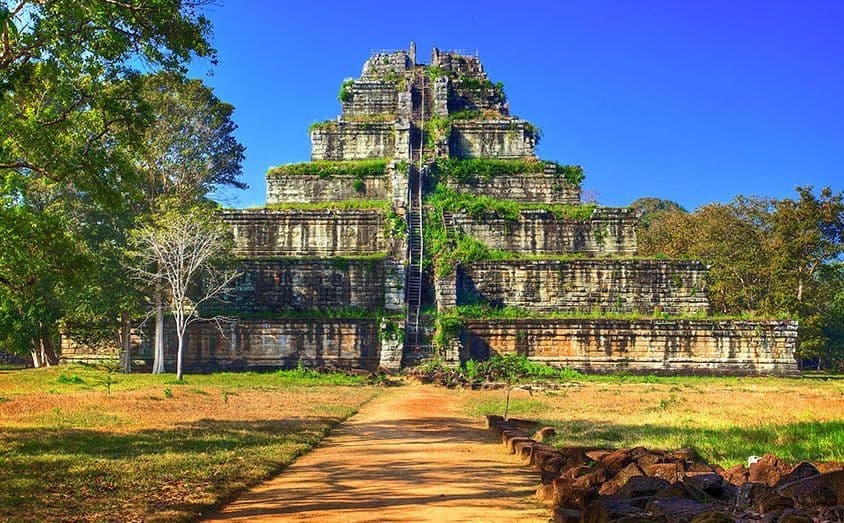 Un grande e antico tempio piramidale in pietra, con vegetazione rigogliosa sui suoi gradoni, visto da un sentiero sterrato sotto un cielo azzurro.