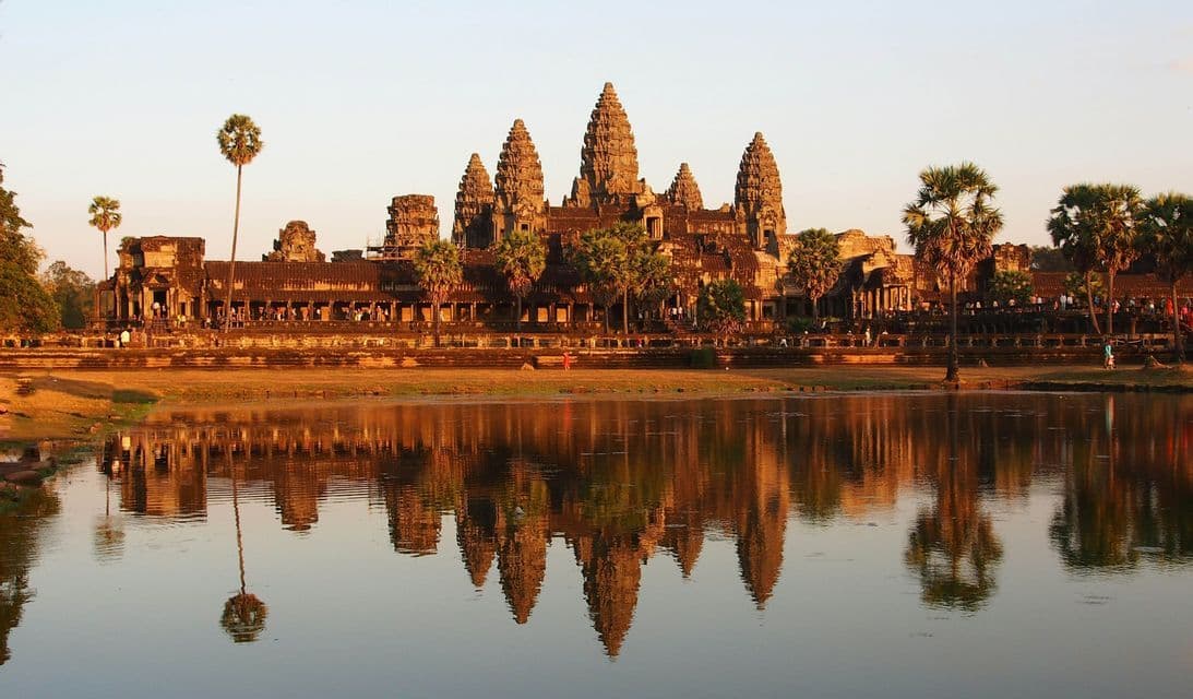 Un antico tempio in pietra con molteplici torri si riflette perfettamente in un grande lago calmo all'alba, circondato da palme.