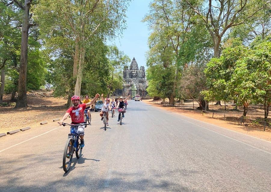 Un gruppo WeRoad sorride pedalando in bicicletta su una strada alberata verso un grande e antico portale in pietra.