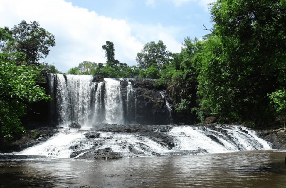Un'ampia cascata si riversa su rocce scure e a gradoni in un fiume calmo, circondata da alberi verdi rigogliosi sotto un cielo parzialmente nuvoloso.