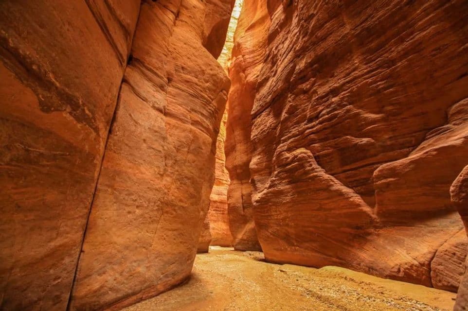 A narrow, sandy path winds through a slot canyon with towering, layered orange rock walls.
