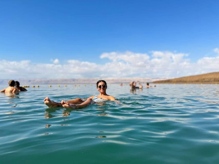 A woman on a WeRoad group trip floats on her back in calm, turquoise water, with other travelers and arid hills in the background.
