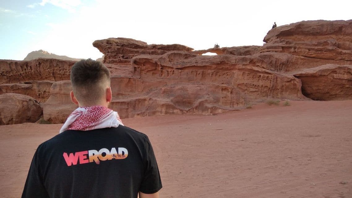A person viewed from behind, wearing a WeRoad t-shirt and keffiyeh, looks out at large red rock formations in a desert.