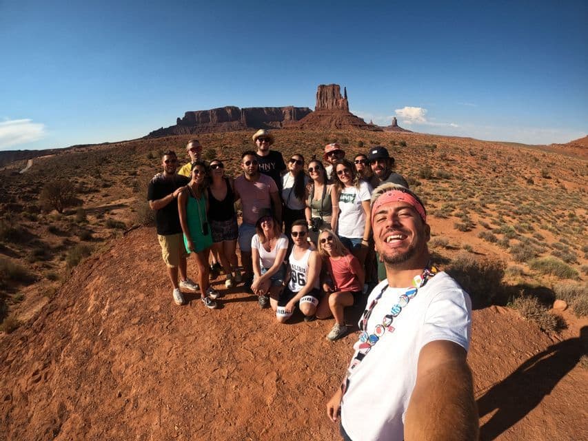 A man takes a selfie with a WeRoad group trip posing on a hill with desert rock formations in the background under a clear sky.
