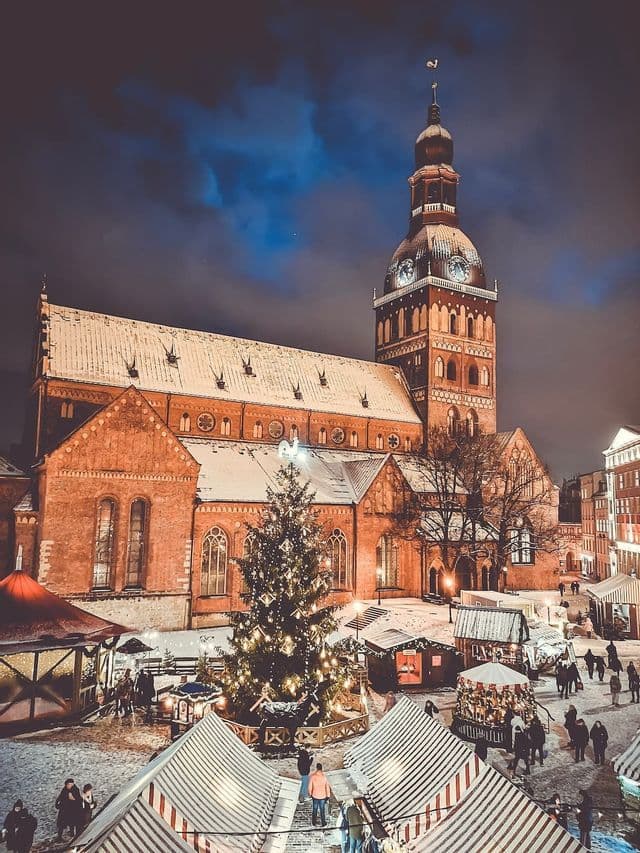 Una piazza innevata di notte ospita un mercatino di Natale con un grande albero decorato, davanti a una cattedrale di mattoni illuminata.
