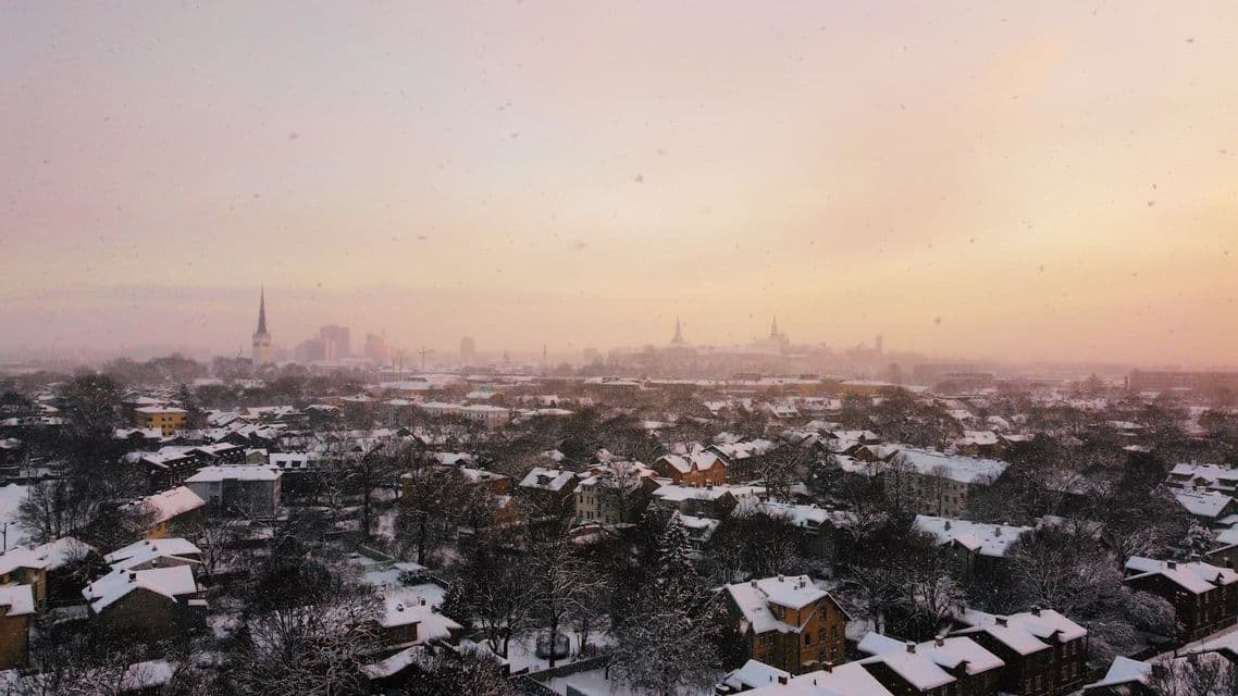 Vue aérienne d'un paysage urbain enneigé avec des toits et des arbres couverts de blanc lors d'un coucher de soleil brumeux aux tons chauds.