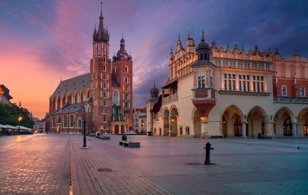 Una plaza de ciudad adoquinada vacía con una alta iglesia de ladrillo y un salón ornamentado bajo un cielo de amanecer colorido.