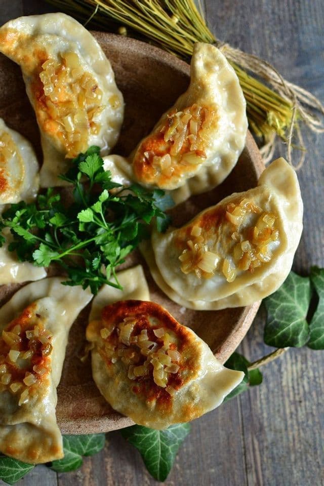 Primer plano de pierogi fritos en sartén sobre plato de madera, con cebolla salteada y perejil fresco.