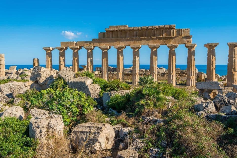 Le rovine di un antico tempio greco con grandi colonne di pietra si ergono su una costa che si affaccia sul mare blu sotto un cielo limpido.