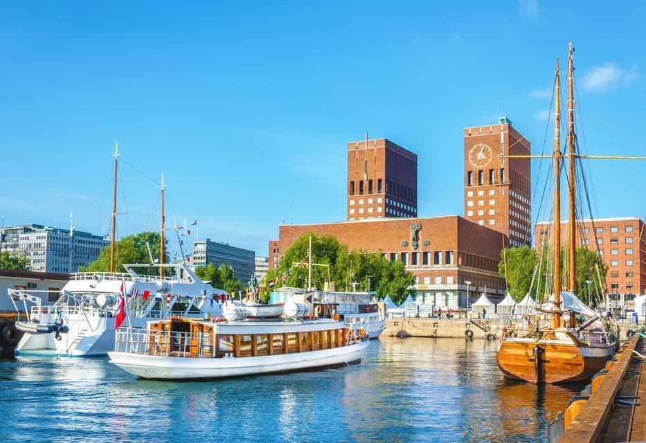 Barche di varie dimensioni ormeggiate in un porto, con un grande edificio in mattoni rossi a doppia torre che si staglia contro un cielo azzurro brillante.