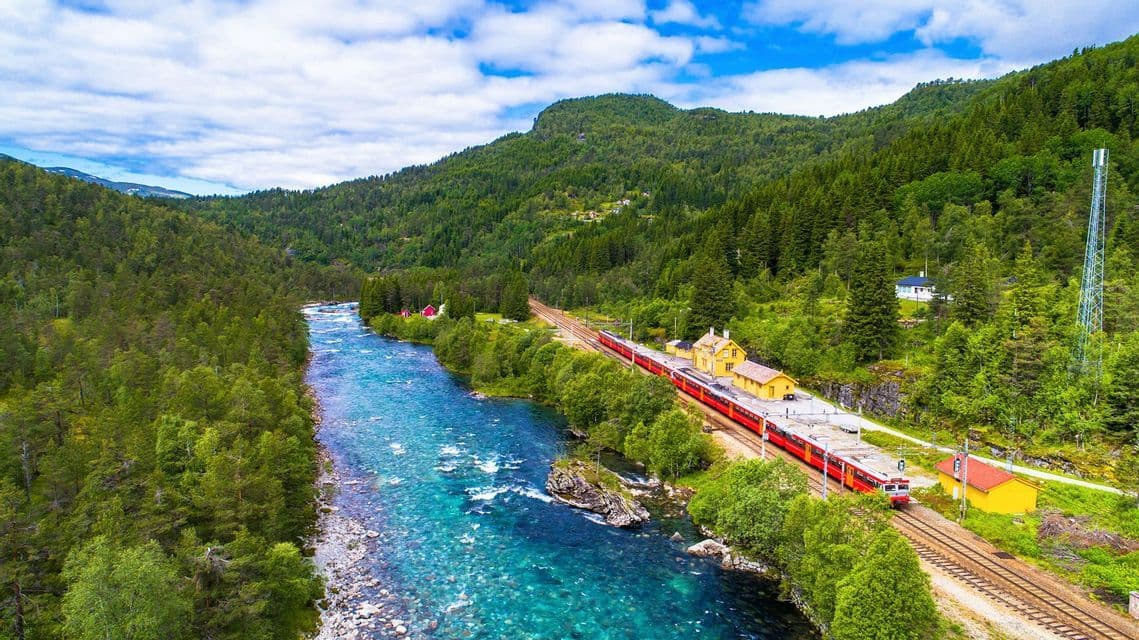 Veduta aerea di un treno rosso fermo in stazione, accanto a un fiume azzurro e cristallino che attraversa una valle di fitte foreste verdi e maestose montagne.