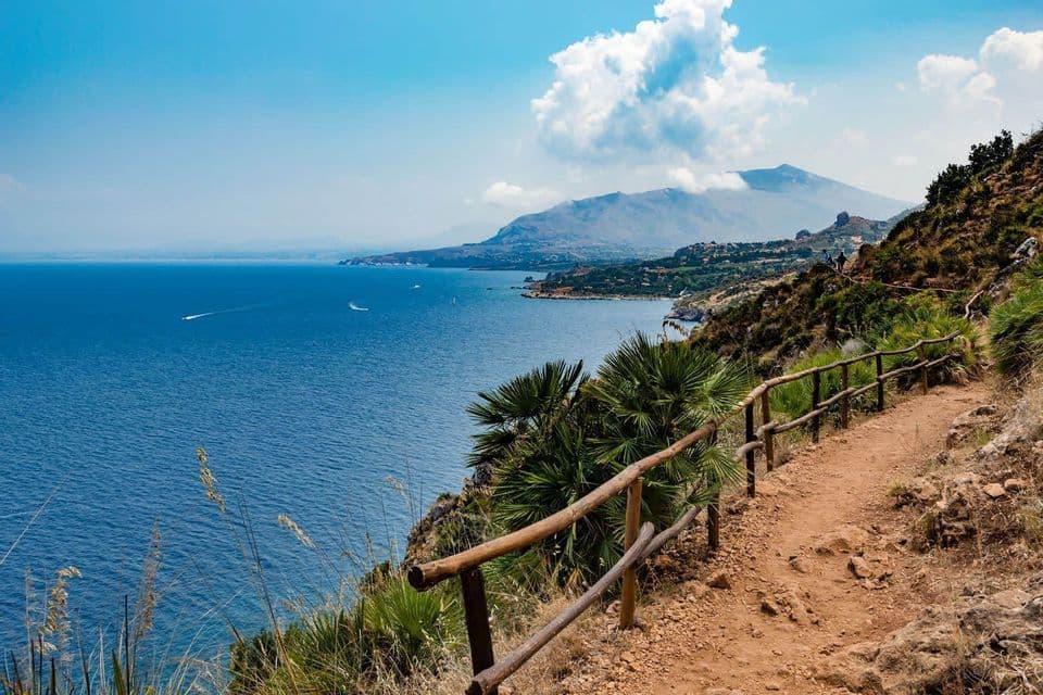 Un chemin de terre avec une clôture en bois longe une falaise côtière surplombant la mer bleue et une chaîne de montagnes lointaine.