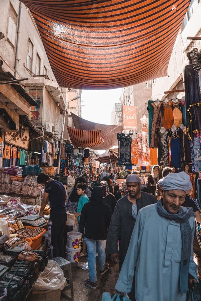 A crowd of people walks through a bustling street market lined with stalls and covered by a striped orange awning.