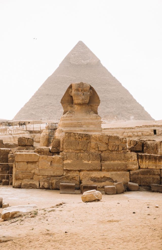 Le Grand Sphinx se dresse devant une grande pyramide dans un paysage sablonneux, avec des ruines de pierre antiques au premier plan.