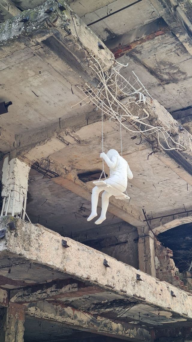 Eine weiße Skulptur einer Person, die den Kopf in den Händen hält, sitzt auf einer Schaukel in einem verfallenden Betongebäude.