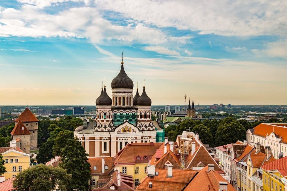 Eine reich verzierte Kathedrale mit schwarzen Zwiebeltürmen erhebt sich über einer historischen Stadtlandschaft mit roten Ziegeldächern und grünen Bäumen unter blauem Himmel.