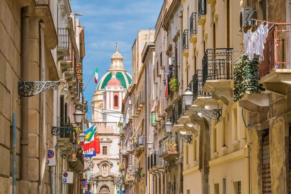 Una stretta via italiana fiancheggiata da edifici e balconi, con una grande cupola di cattedrale visibile in lontananza sotto un cielo blu.