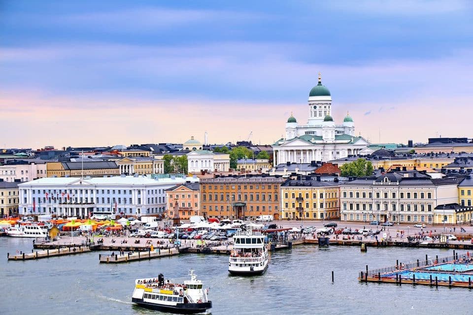Vista della città di un porto trafficato con due traghetti, edifici classici e una grande cattedrale bianca con cupole verdi sullo sfondo.