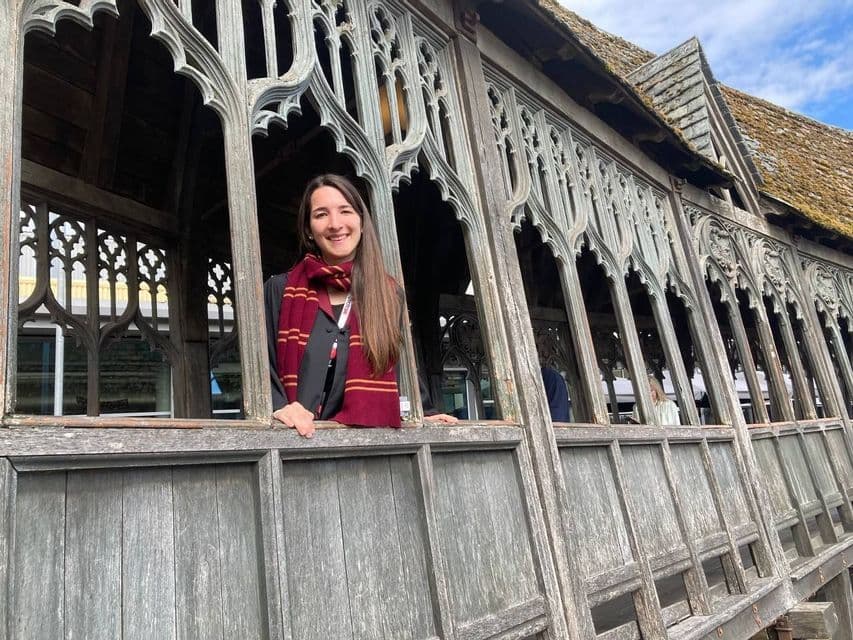 Una donna sorridente, con una tunica nera e una sciarpa rossa e oro, si affaccia da una finestra ad arco su un ballatoio rustico in legno.