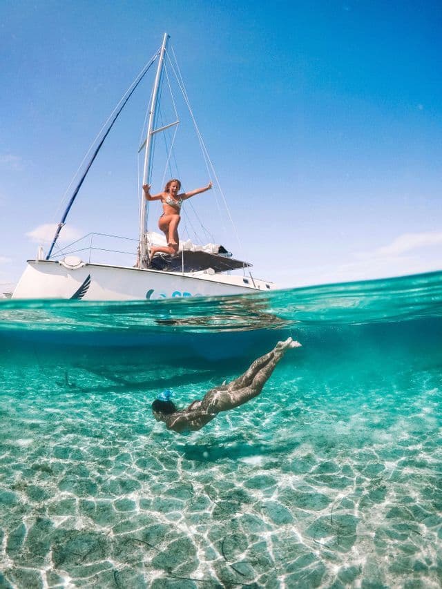 A split-shot of a WeRoad group trip with one woman on a sailboat and another swimming underwater in clear turquoise sea.