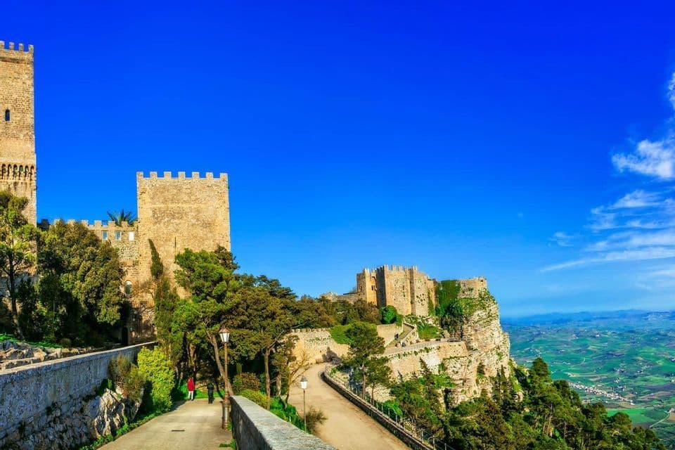 Un castello medievale in pietra con torri, arroccato su un'alta scogliera, che domina una rigogliosa valle verde sotto un cielo blu profondo.
