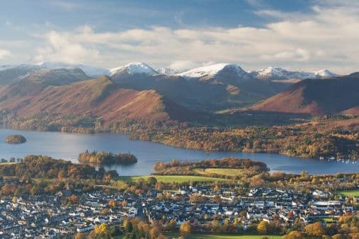 An aerial view of a lakeside town surrounded by autumn trees, with a large lake and snow-capped mountains in the background.