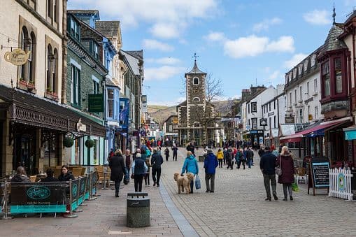 A crowd of people and a dog walk down a busy pedestrian street lined with shops and cafes, with a stone clock tower in the background.