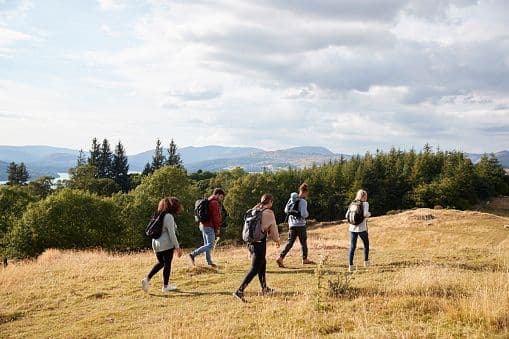 A WeRoad group trip of people with backpacks hiking on a grassy hill with a view of a forest, a lake, and mountains.