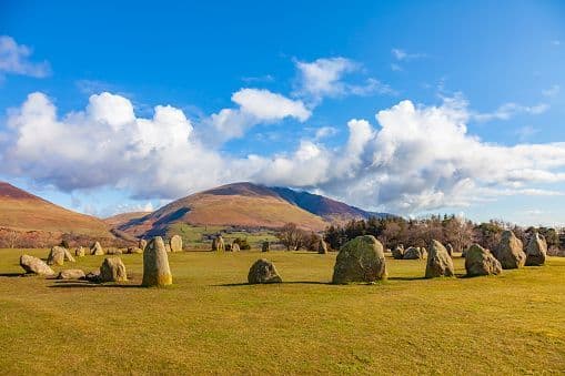A circle of large standing stones rests in a grassy field with rolling hills and a bright blue, cloudy sky in the background.