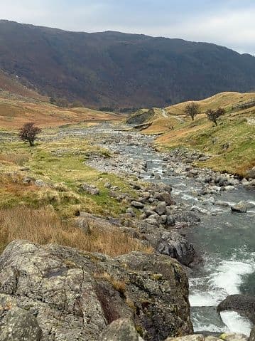 A rocky river flows through a grassy valley at the foot of a large mountain under an overcast sky.