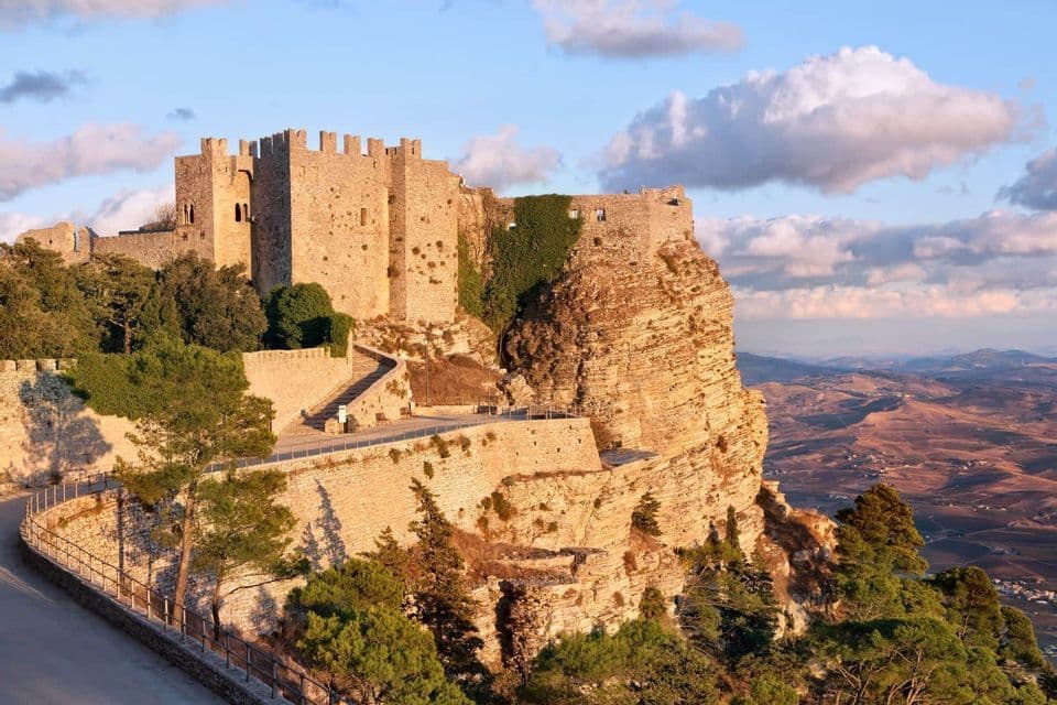 Un château de pierre perché sur une falaise rocheuse surplombant une vallée, illuminé par la lumière chaude du coucher de soleil.