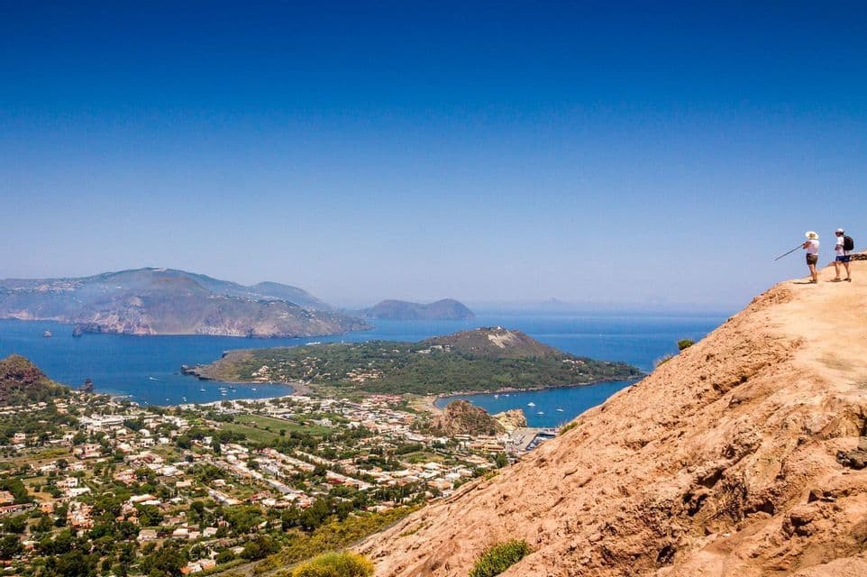 Due persone su una scogliera ammirano una vista panoramica di una città costiera e delle isole nel mare.