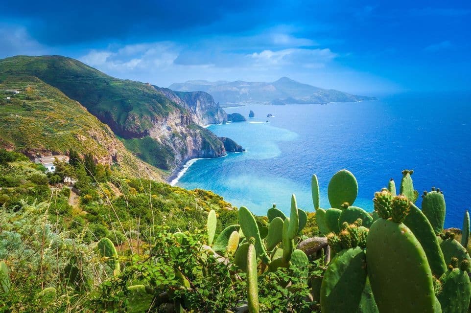 Vista dall'alto su fichi d'India, una costa verde e frastagliata, ripide scogliere e un mare turchese sotto un cielo blu drammatico.