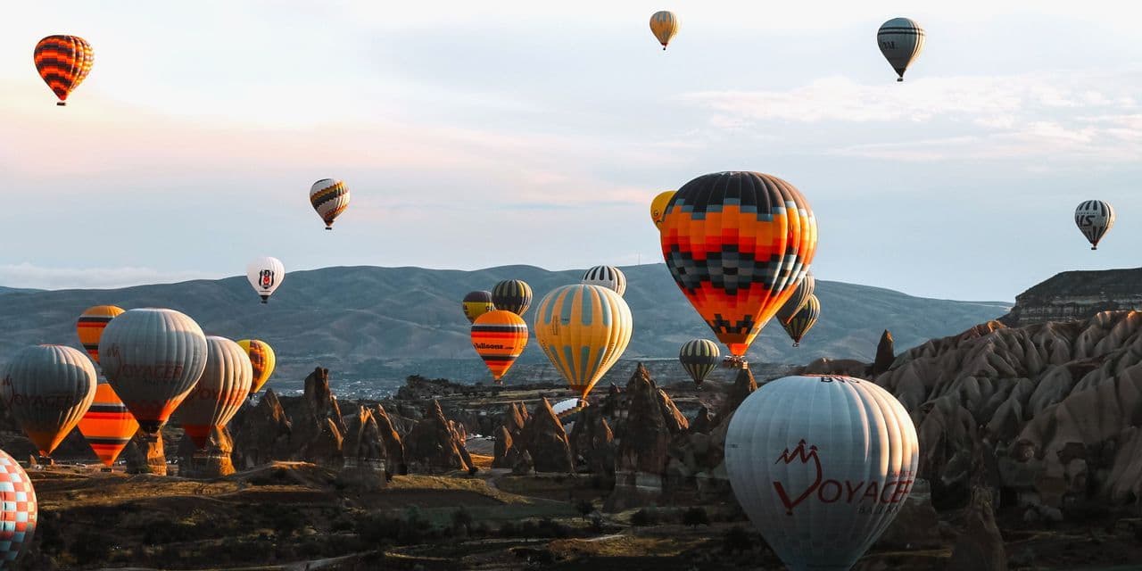 Many colorful hot air balloons flying over a rocky landscape with pointed formations at sunrise.