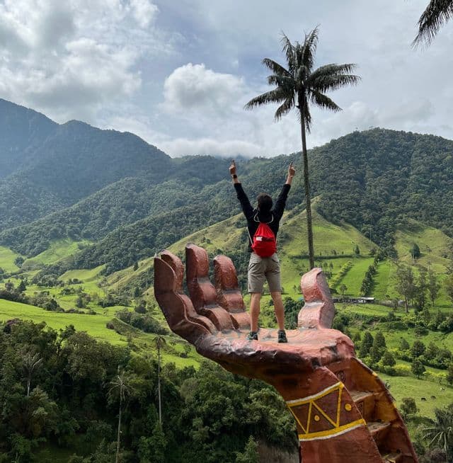 Eine Person mit rotem Rucksack steht auf einer großen Handskulptur mit erhobenen Armen, mit Blick auf ein üppiges grünes Tal und eine hohe Palme.