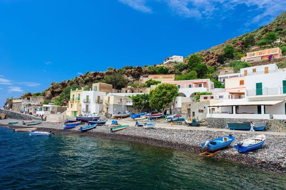 Barche colorate sono ormeggiate su una spiaggia rocciosa in un borgo marinaro adagiato su una collina verde, sotto un cielo azzurro e limpido.