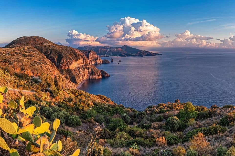 Una costa aspra e assolata con scogliere a picco incontra il mare azzurro, con isole lontane sotto un cielo con grandi nuvole bianche.