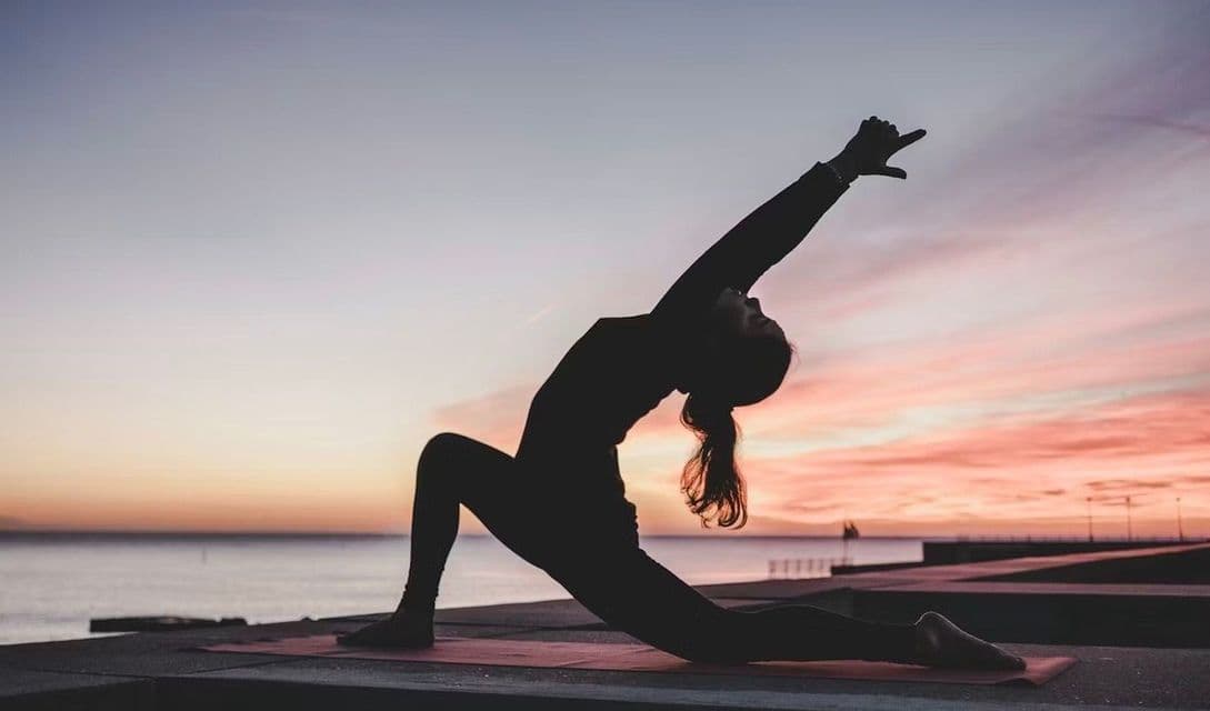 Silhouette di una persona che pratica yoga su un tappetino su un molo, con il mare e un tramonto colorato sullo sfondo.